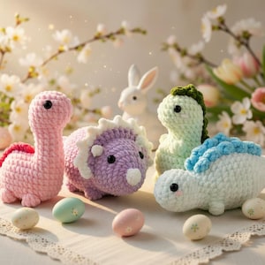 Op de afbeelding: Handgemaakte gehaakte dinosaurussen in roze, paars, groen en wit, met decoratieve eieren. De dinosaurussen hebben zwarte ogen en zijn gerangschikt op een met kant afgewerkt oppervlak, met een wit konijn op de achtergrond.