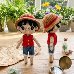 May include: Two crochet dolls of a character wearing a red vest, blue shorts, and a straw hat with a red band. The dolls are standing on a wooden surface with a treasure map, a compass, and small glass bottles with shells.