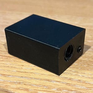May include: A matte black rectangular block with a smooth finish. One side features a circular threaded opening and a small, recessed circular hole positioned to the right of the threaded port. The object has a solid, uniform construction.