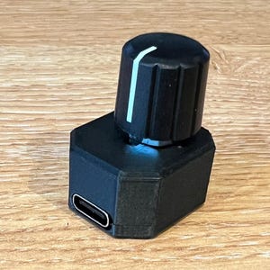 May include: A black electronic device with a rotating knob and a rectangular base. The knob has a white stripe and is cylindrical with vertical grooves. The base has a rectangular port. The device is on a wooden surface.