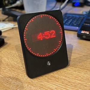 May include: A black rectangular device with a circular red digital display showing the number "4:52". The display is surrounded by a ring of red lights. A small logo is visible on the lower front of the device.