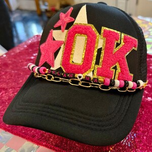 May include: Black trucker hat with a mesh back and a curved brim. The front features the word "OK" in pink, outlined in gold glitter, with a star design. A beaded chain with gold links and letter beads is attached.