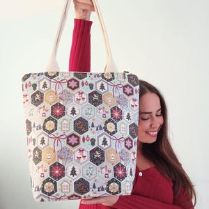 Peut inclure: Un sac cabas blanc avec un motif de Noël représentant un motif répété d'hexagones avec divers éléments de fête, notamment des flocons de neige, des rennes, des sapins de Noël et des ornements. Le sac a des anses de couleur crème.