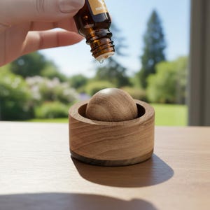 May include: A wooden essential oil diffuser with a spherical top and a circular base. A hand is holding a brown glass bottle, dispensing a drop of oil into the diffuser. The diffuser is made of light brown wood.