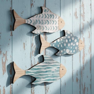 Puede incluir: Tres decoraciones de pared de peces de madera. Cada pez tiene una cola y cabeza de madera clara, con un cuerpo pintado en tonos de azul y blanco. Los cuerpos de los peces presentan diferentes patrones: chevrones, lunares y rayas horizontales. El fondo es una pared de madera azul desgastada.