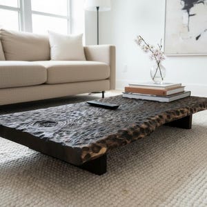 Peut inclure: Une table basse rectangulaire marron foncé avec un bord brut. La table est en bois et repose sur des pieds noirs. Des livres et une télécommande sont sur la table. La table mesure environ 152 cm de long.