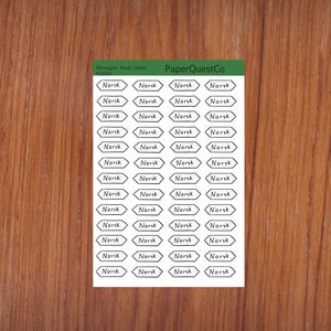 May include: A sheet of white paper with black and white labels that say "Norsk" in a handwritten font. The labels are in the shape of a diamond with rounded corners. The paper is on a woodgrain background.