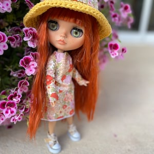 Può includere: Una bambola dai capelli rossi con un vestito floreale e un cappello all'uncinetto giallo con un bordo floreale. La bambola è in piedi davanti a un cespuglio di fiori rosa.