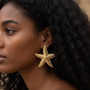 May include: Gold-colored starfish earrings. The earrings are large and textured, with a detailed design mimicking the natural form of a starfish. The earrings are worn on a person's ear, adding a touch of coastal charm.