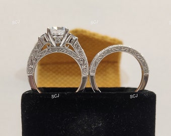 May include: A pair of silver engagement rings with intricate designs. The engagement ring features a large, round, clear center stone with two smaller stones on either side. The wedding band is a simple band with a delicate design.