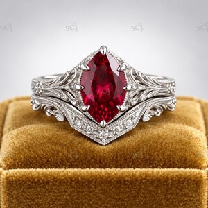 Puede incluir: Un anillo de tono plateado con una gran piedra preciosa roja de corte marquesa en el centro. La banda presenta un intrincado trabajo de metal y detalles de filigrana, complementado por una banda curva con diamantes engastados en pavé.