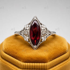 Puede incluir: Un anillo de plata con una gema roja oscura con corte marquesa. El anillo presenta un diseño de filigrana ornamentado con motivos de hojas y corazones. El anillo está sobre una superficie de terciopelo dorado.