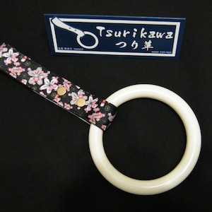 Puede incluir: Asa de anillo blanca con una correa de tela floral negra y rosa. La correa tiene detalles dorados y el texto "Tsurikawa つり革 MADE FOR RIDE" está sobre un fondo azul.