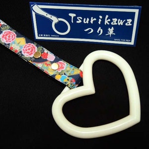 Puede incluir: Manija de coche en forma de corazón blanca con una correa de patrón floral negro. La correa tiene detalles dorados y el texto "Tsurikawa つり革" está en una etiqueta azul.