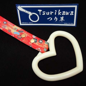 Puede incluir: Asa de coche en forma de corazón blanca con una correa estampada en rojo, amarillo y azul con botones dorados. La correa tiene el texto "Tsurikawa つり革" y "Made for Ride".