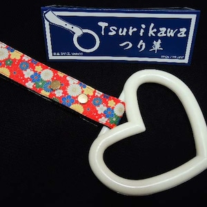 Puede incluir: Mango de coche blanco en forma de corazón con una correa de patrón floral rojo, blanco y azul. La correa tiene dos broches metálicos. El mango está unido a una caja azul y blanca con el texto "Tsurikawa つり革" impreso en ella.