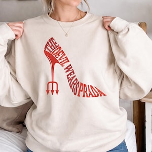 Puede incluir: Sudadera de color crema con un diseño de zapato de tacón alto rojo. El zapato tiene forma de tridente e incorpora el texto "THE DEVIL WEARS PRADA" en una fuente estilizada. La sudadera tiene mangas largas.