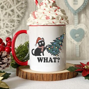 May include: White ceramic mug with a red handle. The mug has a black cat wearing a Santa hat and a Christmas tree with colorful ornaments. The text "WHAT?" is printed on the mug.