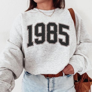 May include: Gray sweatshirt with a black and brown leopard print 1985 design.