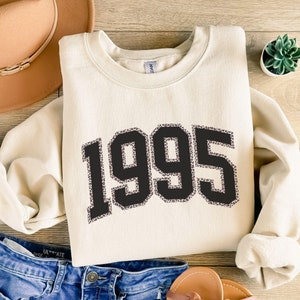 May include: A cream-colored sweatshirt with a black and white leopard print design featuring the year "1995" in large, bold letters.