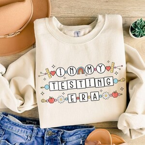 May include: A beige sweatshirt with the text "In My Testing Era" in colorful block letters. The text is surrounded by a chain of colorful school supplies, including a rainbow, a heart, a ruler, a pencil, a daisy, and an apple.