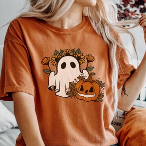 K&ouml;nnte beinhalten: Orangefarbenes T-Shirt mit kurzen &Auml;rmeln, das einen Cartoon-Geist mit Sonnenblumenkranz, K&uuml;rbis und Herbstlaub zeigt. Das Shirt besteht aus weichem Material und ist perfekt f&uuml;r Halloween oder den Herbst. Das Model tr&auml;gt eine dunkelblaue Jeans.