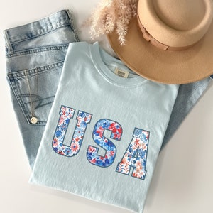 Op de afbeelding: Een lichtblauw T-shirt met het woord "USA" erop gedrukt. De letters zijn gemaakt van een bloemenpatroon in rood, wit en blauw.