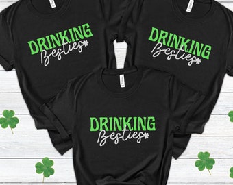 Puede incluir: Tres camisetas negras con el texto "Drinking Besties" en letras verdes y blancas. Las camisetas están dispuestas sobre una superficie de madera blanca.