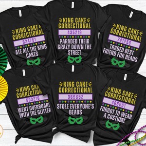 May include: Six black t-shirts with a Mardi Gras theme. Each shirt features the text "King Cake Correctional" with a different number and a funny phrase. The shirts are laid out on a wooden surface with Mardi Gras decorations.