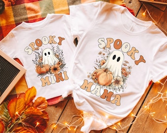 Może przedstawiać: Białe koszulki z motywem Halloween. Jedna koszulka z napisem "Spooky Mini", druga "Spooky Mama". Każda z nich przedstawia ducha, dynie i kwiaty w stylu retro, idealne na święto.