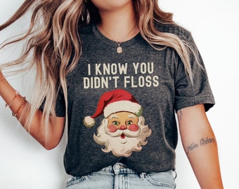 Op de afbeelding: Donkergrijs t-shirt met de tekst "I KNOW YOU DIDN'T FLOSS" en een vintage afbeelding van de Kerstman. De Kerstman heeft een rode muts, een witte baard en roze wangen. Een casual, feestelijk ontwerp.