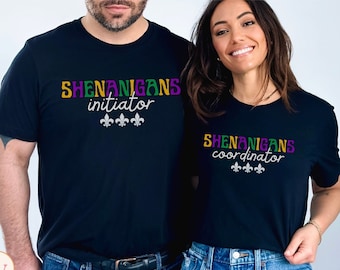 May include: Two black t-shirts with a Mardi Gras theme. The text on the shirts reads "Shenanigans initiator" and "Shenanigans coordinator". Both shirts feature three fleur-de-lis.