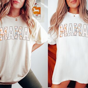 May include: Two women wearing white t-shirts with the word "MAMA" printed in large, bold letters. The letters are filled with a floral pattern in shades of pink, orange, and blue. The shirts are a relaxed fit and have short sleeves.