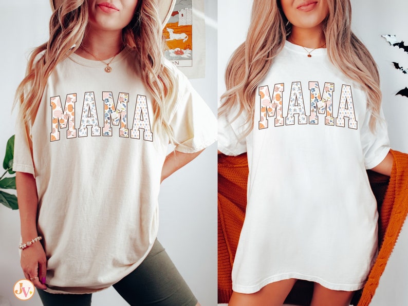 May include: Two women wearing white t-shirts with the word "MAMA" printed in large, bold letters. The letters are filled with a floral pattern in shades of pink, orange, and blue. The shirts are a relaxed fit and have short sleeves.