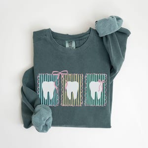 May include: A teal-colored sweatshirt featuring three cartoon teeth in rectangular frames. Each tooth has a pink bow or dental floss. The frames have a striped background with a scalloped border. The text "BLUE SPRUCE" is at the bottom left.