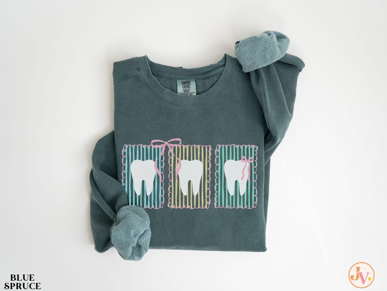 May include: A teal-colored sweatshirt featuring three cartoon teeth in rectangular frames. Each tooth has a pink bow or dental floss. The frames have a striped background with a scalloped border. The text "BLUE SPRUCE" is at the bottom left.