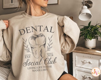 May include: A beige sweatshirt with the text "Dental Social Club" and a drawing of a tooth. The text also includes "Est. 2024" and "Living life to the maxillary."
