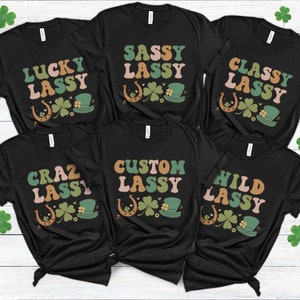 May include: Black t-shirts with various text designs including "Lucky Lassy," "Sassy Lassy," and "Classy Lassy." Each shirt features a horseshoe, shamrock, and a green hat graphic. The shirts are decorated with retro-style fonts and colors.