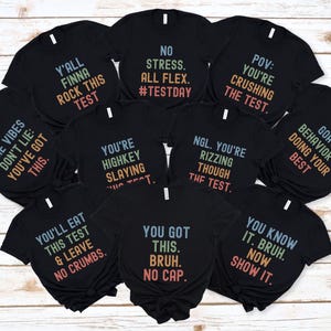 May include: Nine black t-shirts with different motivational phrases printed on them in a rainbow gradient. The phrases include "Y'all Finna Rock This Test", "The Vibes Don't Lie: You've Got This", "No Stress. All Flex. #TestDay", "You're Highkey Slaying This Test", "POV: You're Crushing The Test", "NGL. You're Rizzing Though The Test", "You'll Eat This Test & Leave No Crumbs", "You Got This. Bruh. No Cap", "Goat Behavior = Doing Your Best", and "You Know It. Bruh. Now Show It."