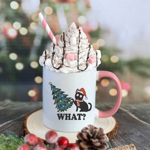 May include: White ceramic mug with a pink handle, featuring a black cat wearing a Santa hat and a Christmas tree. The mug is filled with hot chocolate topped with whipped cream, chocolate drizzle, and pink marshmallows. The text "WHAT?" is printed on the mug.