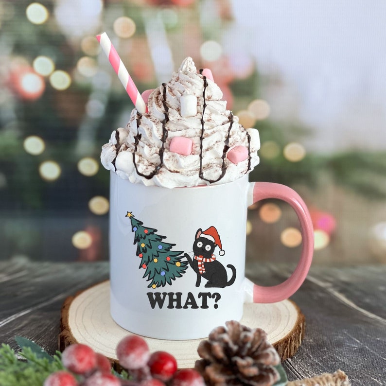 May include: White ceramic mug with a pink handle, featuring a black cat wearing a Santa hat and a Christmas tree. The mug is filled with hot chocolate topped with whipped cream, chocolate drizzle, and pink marshmallows. The text "WHAT?" is printed on the mug.