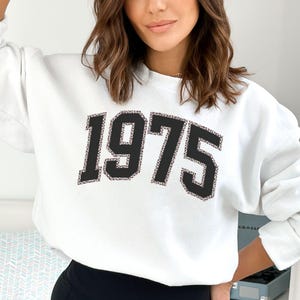 May include: White sweatshirt with a black 1975 graphic on the front.