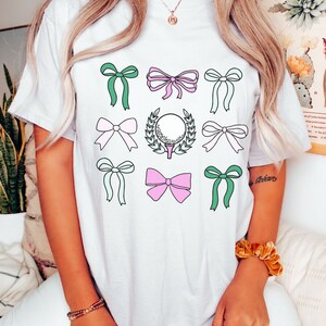 May include: White t-shirt featuring a grid of colorful bows in green, pink, and light blue. A golf ball on a tee with a laurel wreath is in the center. The shirt is a casual, everyday wear item.