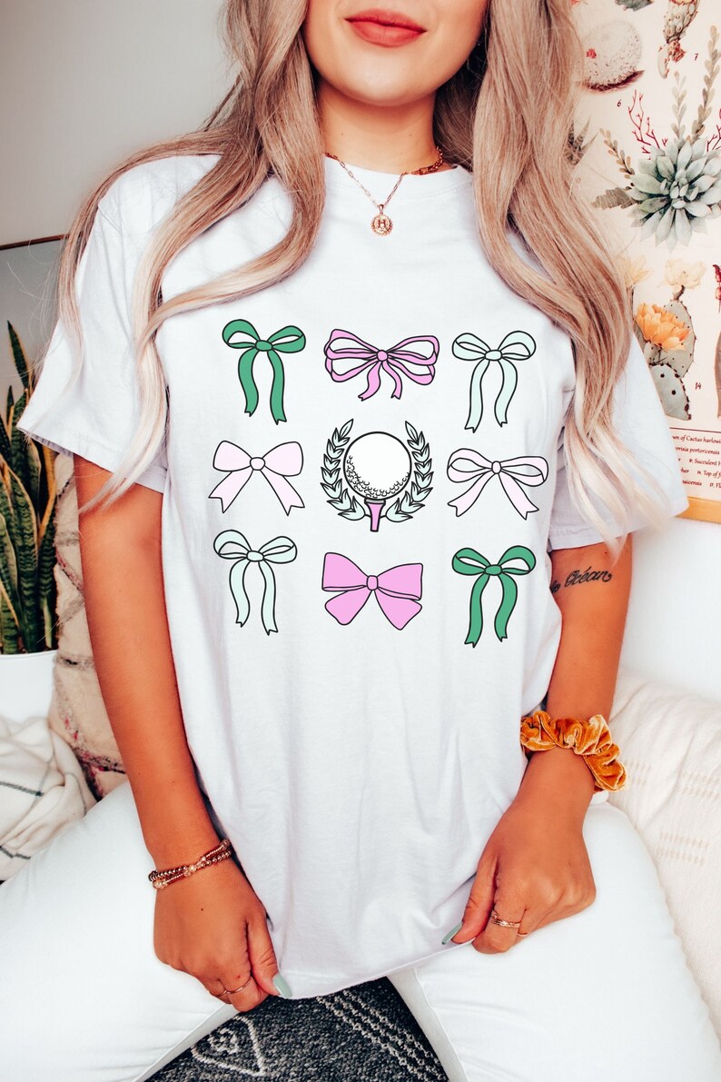 May include: White t-shirt featuring a grid of colorful bows in green, pink, and light blue. A golf ball on a tee with a laurel wreath is in the center. The shirt is a casual, everyday wear item.
