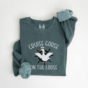 Puede incluir: Una sudadera azul verdosa con el texto "Cruise Goose on the Loose" y un dibujo animado de un ganso con un sombrero de capit&aacute;n y un volante.