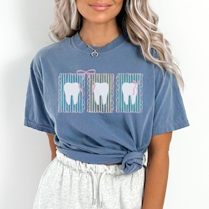 May include: Blue jean t-shirt featuring three cartoon teeth with pink bows. The teeth are set against a striped background. The shirt is a solid blue color and has a relaxed fit.