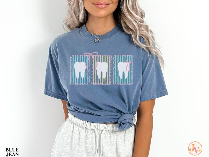 May include: Blue jean t-shirt featuring three cartoon teeth with pink bows. The teeth are set against a striped background. The shirt is a solid blue color and has a relaxed fit.