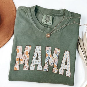 May include: A green t-shirt with the word "MAMA" in large, white letters. The letters are decorated with a patchwork pattern of floral and pumpkin designs.