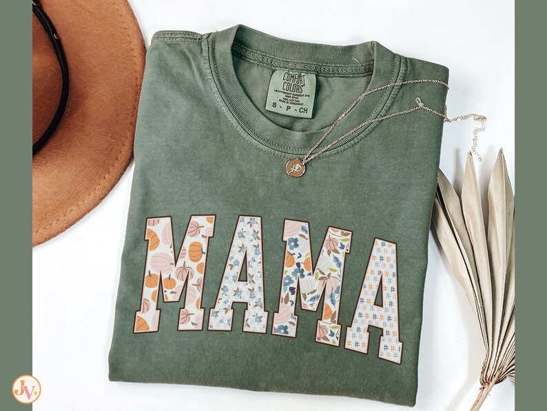 May include: A green t-shirt with the word "MAMA" in large, white letters. The letters are decorated with a patchwork pattern of floral and pumpkin designs.