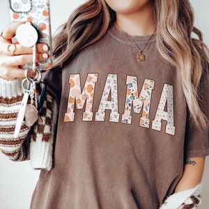 May include: A brown t-shirt with the word "MAMA" printed in large, white letters. The letters are decorated with a floral pattern in shades of pink, blue, and orange.